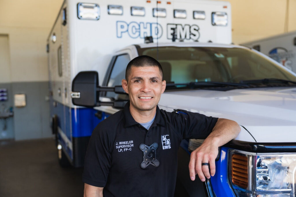 A man in a dark-colored uniform stands with his arm resting on the hood of a white ambulance.