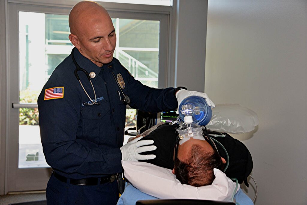 A uniformed EMS provider uses a bag‑valve mask to ventilate a patient lying on a stretcher, with monitoring equipment visible in the background.
