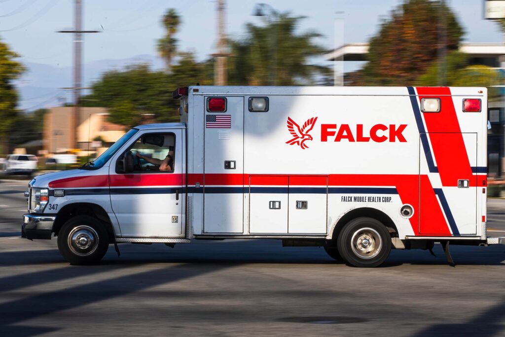 A white and red Falck ambulance with the American flag emblem driving through a suburban intersection in California.